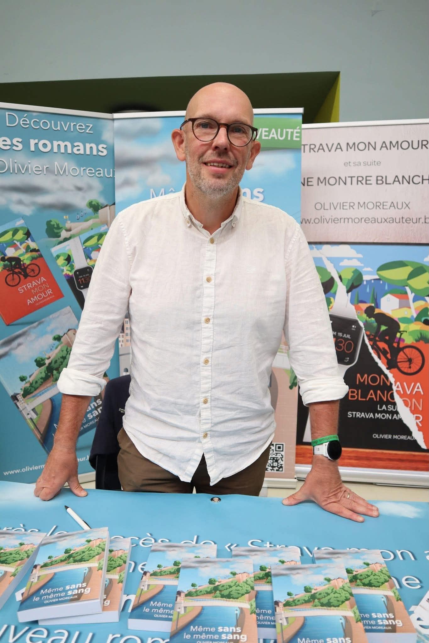 Olivier Moreaux, chemise blanche, debout derrière sa table de dédicaces, entouré de ses affiches et d'exemplaires de « Même sans le même sang »